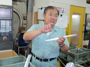 アルミ押し出し材の説明をする大橋さん。同社にはアルミ材は2メートルくらいの長さで搬入されますそれを所定の寸法に切りそろえ、機械にのせ肉を削り、そのあとプレスで穴を掘っていきます。