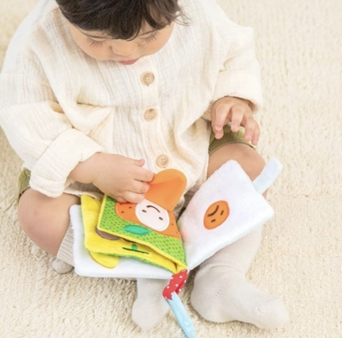 赤ちゃんがお座りしながら遊ぶ様子「ベビー向けの人気商品、布絵本の『もぐもぐばあ』🍊」