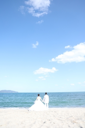 広がる青空と鳥取の砂浜を背景に、手をつなぐ新郎新婦の後ろ姿「U PLUS 米子」